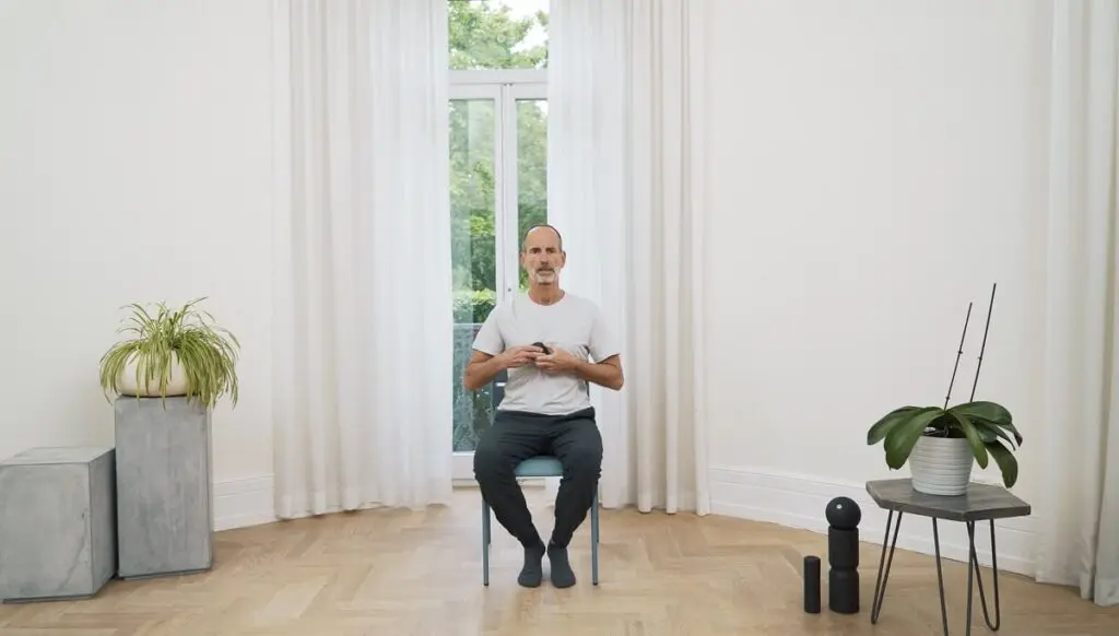 Ein Mann sitzt auf einem Stuhl vor einem Fenster und hält einen Faszienball mit beiden Händen auf das Brustbein gedrückt