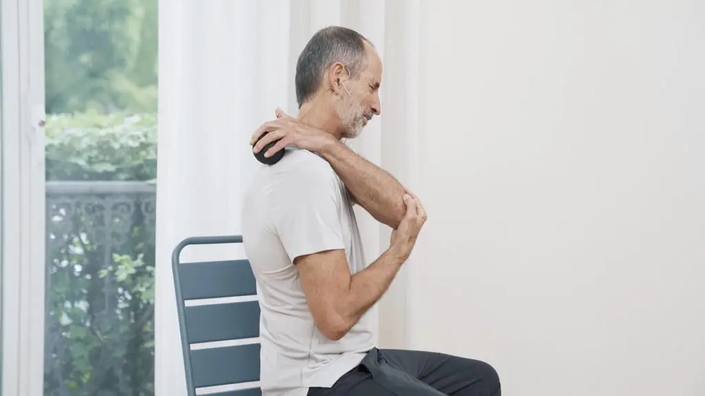 Ein Mann sitzt auf einem Stuhl und hält mit einer Hand eine Faszienkugel, mit der er sich am Schulterblatt massiert