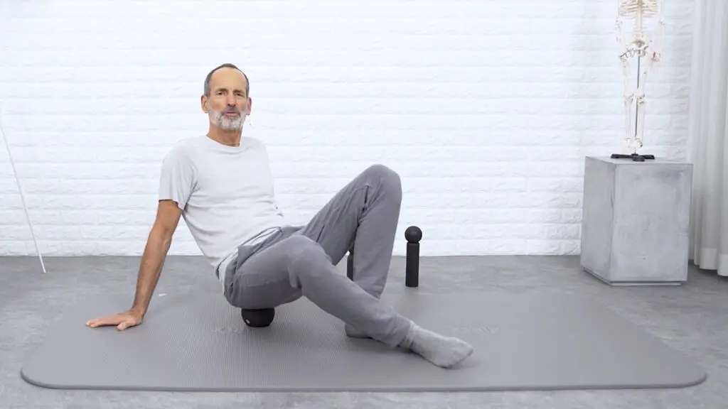 Ein Mann sitzt auf einer mittelgroßen Faszienkugel, stützt sich mit den Händen ab und blickt in die Kamera