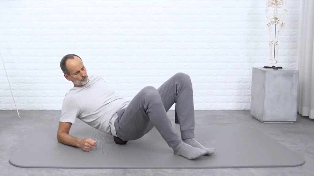 Ein Mann rollt mit dem Gesäß über eine mittelgroße Faszienkugel und stützt sich für die Balance mit den Armen ab