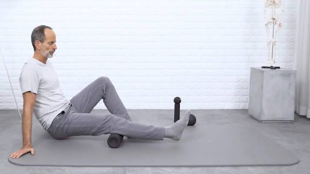 Ein Mann sitzt auf einer grauen Übungsmatte, stützt sich mit den Händen ab und legt sein Bein auf eine mittelgroße Faszienrolle, um eine Faszien-Rollmassage am hinteren Oberschenkel durchzuführen