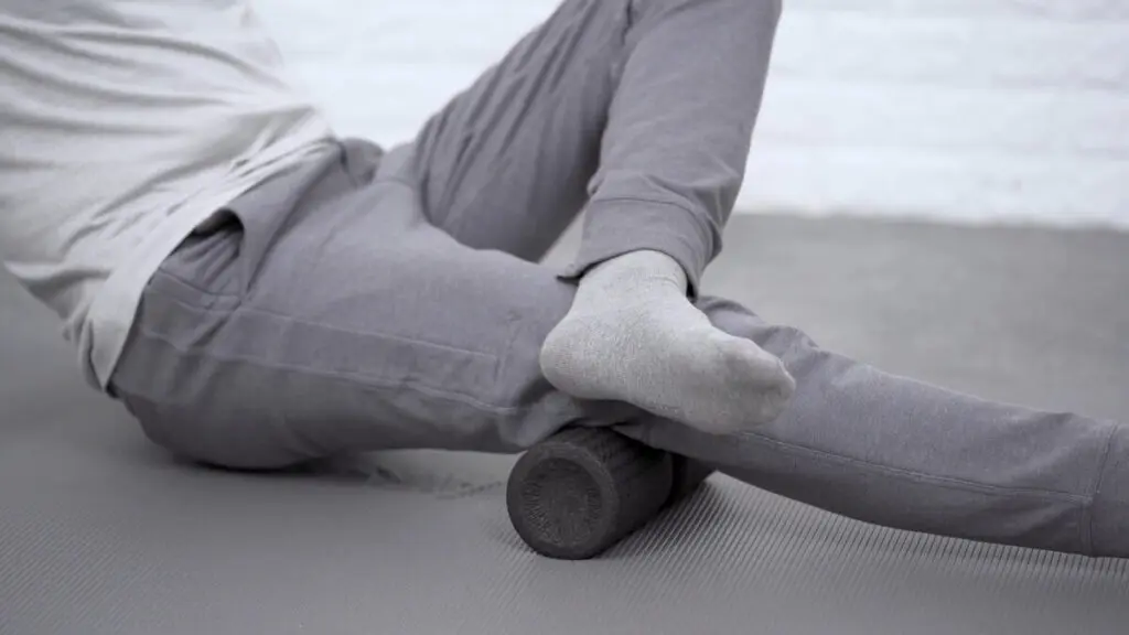 Zu sehen ist, wie ein Mann auf einer grauen Übungsmatte sitzt und seinen hinteren Oberschenkel über eine Faszienrolle rollt. Den Druck intensiviert er, indem er das andere Bein über das abzurollende Bein schlägt.