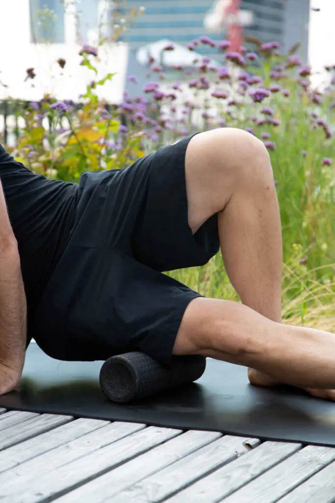 Zu sehen ist ein Mann, der sich mit dem äußeren Oberschenkel auf eine Faszienrolle stützt und so in der Natur eine Faszien-Rollmassage am Oberschenkel durchführt