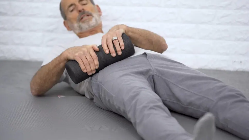 Ein Mann liegt auf einer grauen Übungsmatte und hält in beiden Händen eine Faszienrolle, mit der er den Oberschenkel abrollt