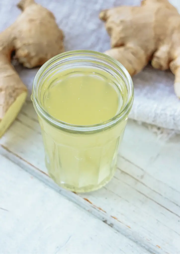 In einem Glas befindet sich eine gelbe Flüssigkeit, daneben liegen Ingwerknollen.