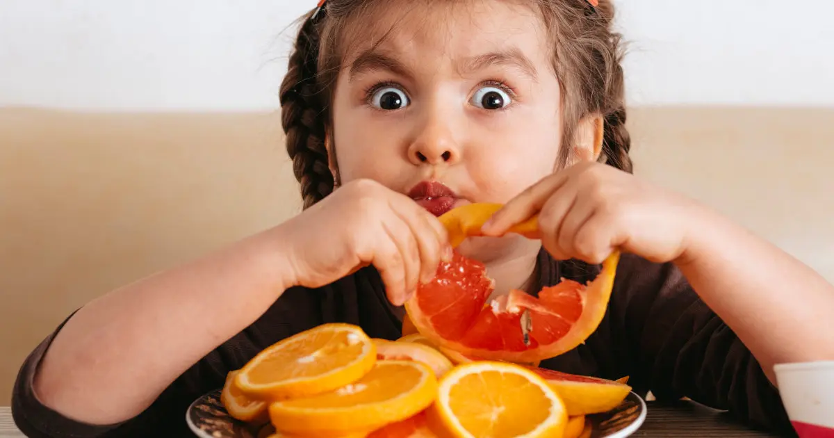Ein Kind sitzt vor einem großen Teller Orangenscheiben und hat gerade beherzt in eine hineingebissen. Die Augen sind groß.