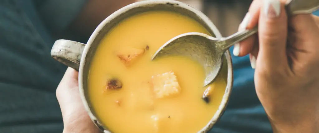 Eine Hand hat eine Schüssel Suppe in der Hand, die andere Hand hält einen Löffel in die Suppe.