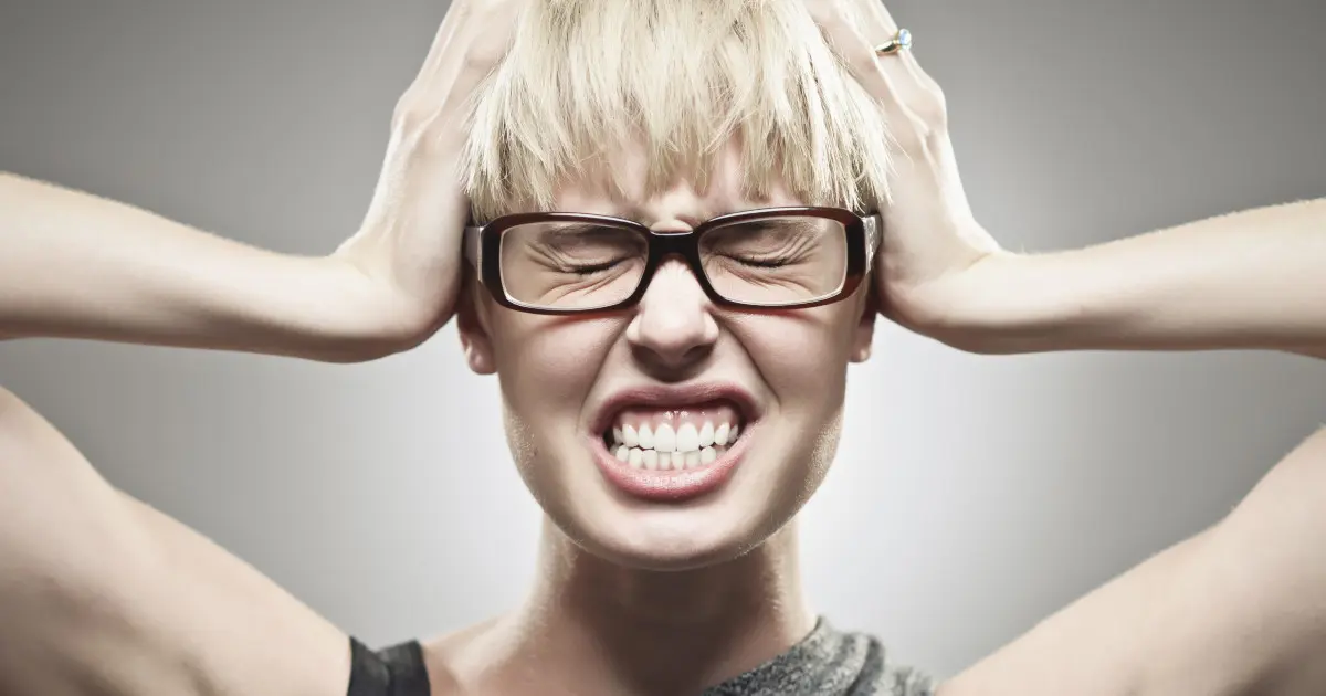 Eine Frau fasst sich mit beiden Händen an ihren Kopf und beißt ihre Zähne fest aufeinander. Die Augen sind geschlossen.