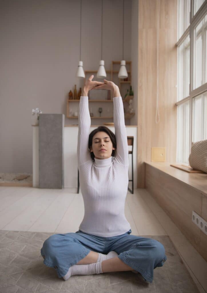 Eine Frau sitzt im Schneidersitz auf dem Boden und streckt beide Arme nach oben.