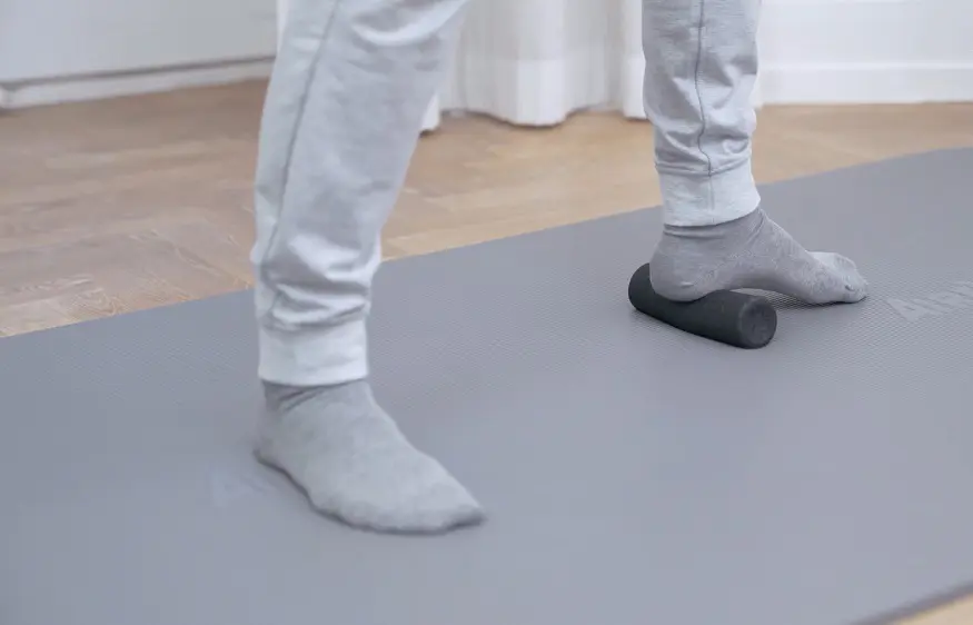 Der linke Fuß steht mit der Ferse auf einer kleinen Faszienrolle, der rechte Fuß stützt seitlich ab
