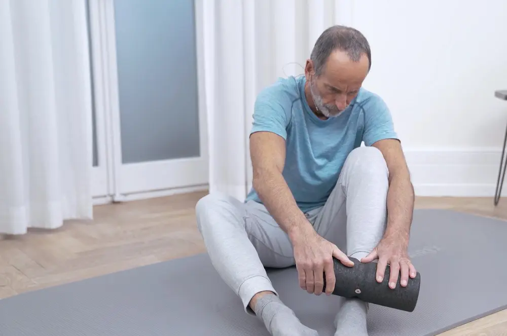 Ein Mann sitzt auf der Übungsmatte und führt mit beiden Händen eine mittelgroße Faszienrolle zur Selbstmassage über das Schienbein