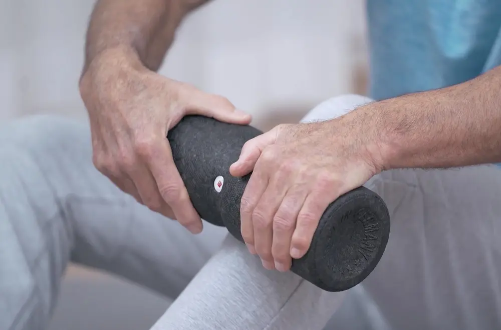 Ein Mann sitzt auf der Übungsmatte und führt mit beiden Händen eine mittelgroße Faszienrolle zur Selbstmassage über das Schienbein