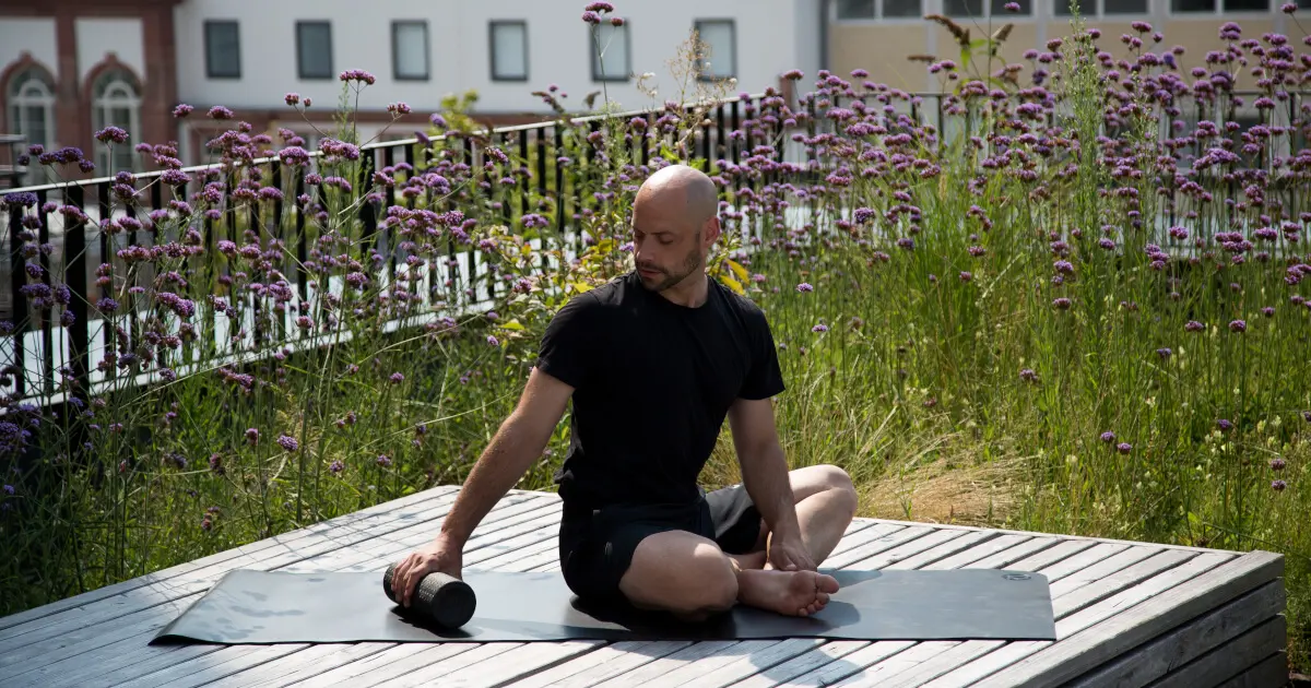Zu sehen ist ein Mann, der auf einer Holzterasse vor einer Blumenwiese mit Blick auf die Stadt sitzt. Hinter ihm befindet sich eine Faszienrolle, er dreht sich zu dieser um und hält sie in einer Hand.