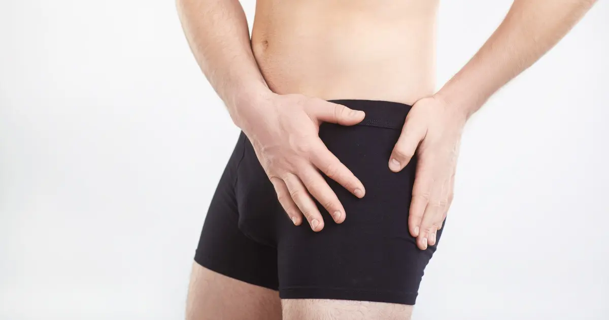 Zu sehen ist ein Mann, der sich mit beiden Händen an die Hüfte fasst. Eine Hand liegt auf der Außenseite über dem großen Rollhügel (Trochanter major) und die andere Hand nahe der Leiste. An diesen Stellen findet sich oft die Ursache bei einer Schleimbeutelentzündung in der Hüfte.