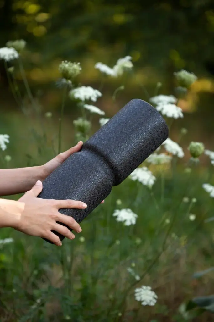 Zu sehen ist eine Hand, die eine Faszienrolle mit Wirbelsäulenvertiefung in die Kamera hält. Im Hintergrund ist eine Blumenwiese mit weißen Blumen zu sehen.