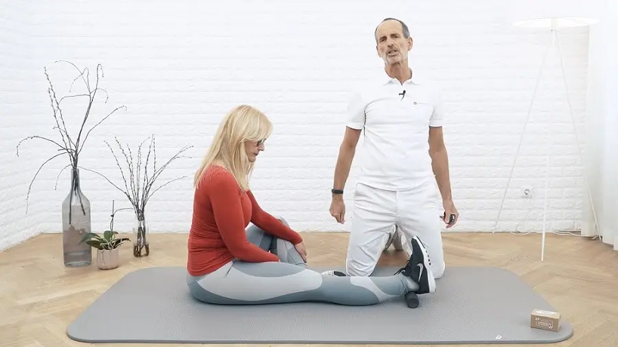 Ein weiß gekleideter Mann kniet neben einer Frau mit rotem Oberteil, die ihr Bein für eine Dehnübung ausgestreckt hat. Unter ihrem Fuß liegt die Mini-Rolle.