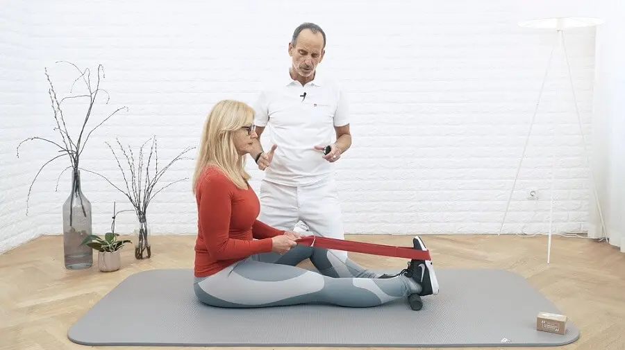 Ein weiß gekleideter Mann kniet neben einer Frau mit rotem Oberteil, die ihr Bein für eine Dehnübung ausgestreckt hat und es mit einer Übungsschlaufe von Liebscher & Bracht dehnt. Unter ihrem Fuß liegt die Mini-Rolle.