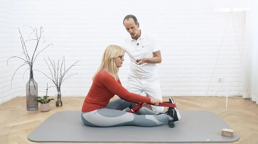Ein weiß gekleideter Mann kniet neben einer Frau mit rotem Oberteil, die ihr Bein für eine Dehnübung ausgestreckt hat und es mit einer Übungsschlaufe von Liebscher & Bracht dehnt. Unter ihrem Fuß liegt die Mini-Rolle.