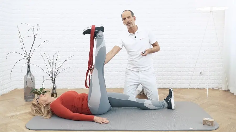 Ein weiß gekleideter Mann kniet neben einer Frau mit rotem Oberteil, die ihr Bein in die Höhe reckt, um eine Dehnübung an der Wade gegen Knieschmerzen durchzuführen