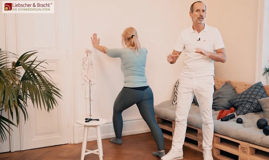 Zu sehen ist eine Frau in einem türkisfarbenen Oberteil, die im Ausfallschritt an eine Wand lehnt, um eine Dehnübung für die Wade durchzuführen. Neben ihr steht ein Mann in weißer Kleidung, der die Übung erklärt.