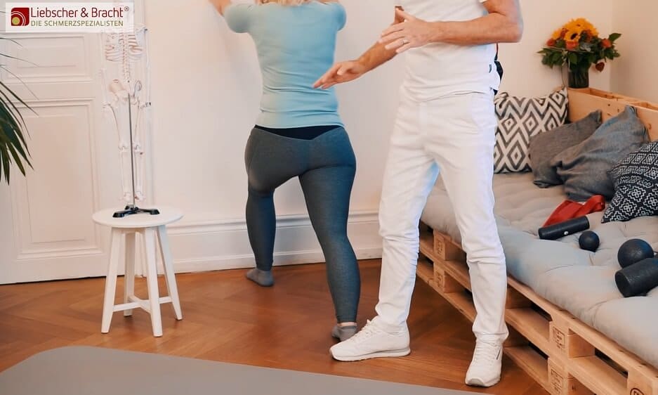 Zu sehen ist eine Frau in einem türkisfarbenen Oberteil, die im nah gezeigten Ausfallschritt an eine Wand lehnt, um eine Dehnübung für die Wade durchzuführen. Neben ihr steht ein Mann in weißer Kleidung, der die Übung erklärt.