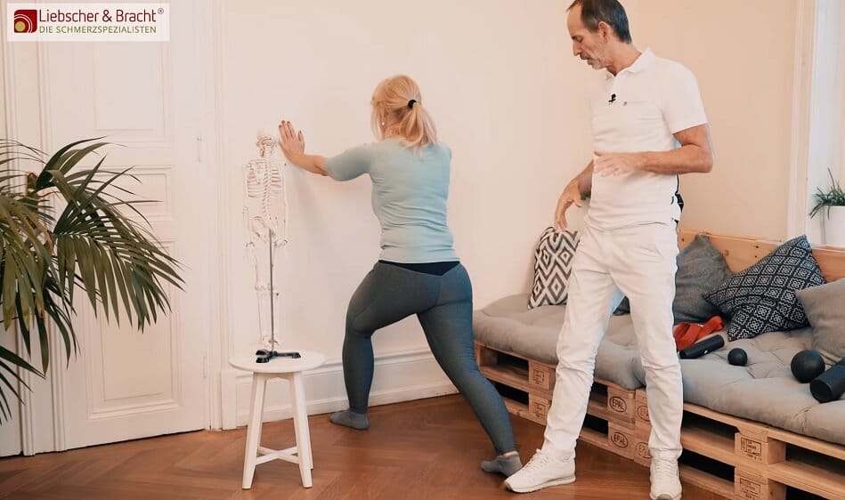 Zu sehen ist eine Frau in einem türkisfarbenen Oberteil, die im Ausfallschritt und mit leicht gebeugtem rechten Knie an eine Wand lehnt, um eine Dehnübung für die Wade durchzuführen. Neben ihr steht ein Mann in weißer Kleidung, der die Übung erklärt.