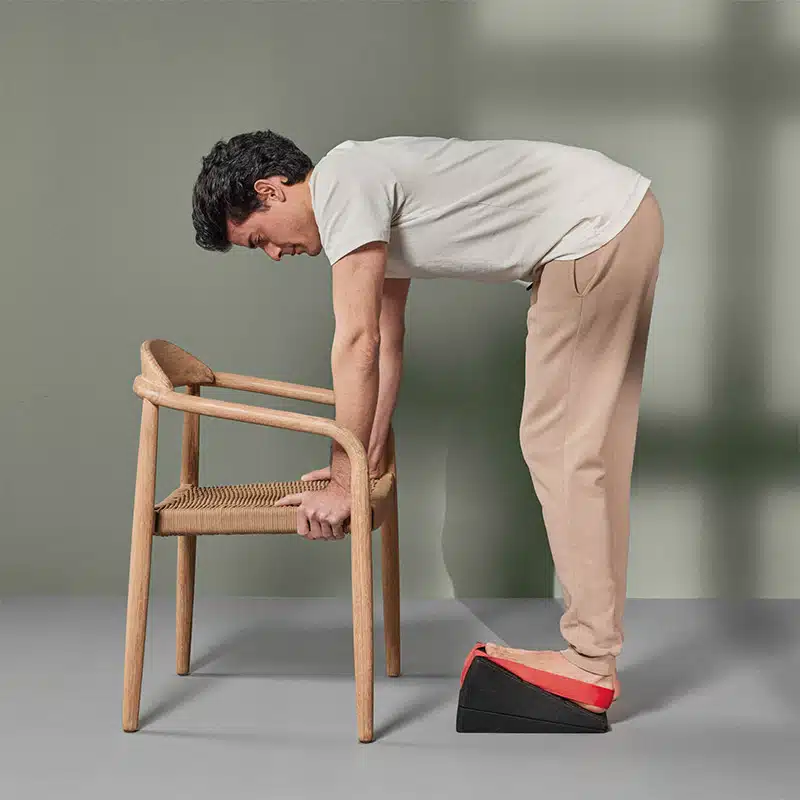 Ein junger Mann in weißem T-Shirt und beiger Baumwoll-Hose steht mit seinem linken Bein auf dem Knieretter. Der Fuß steht in der roten Trittschlaufe, damit er nicht abrutscht. Er beugt sich mit geradem Rücken nach unten, um sich an den Seiten der Sitzfläche eines Holzstuhls abzustützen.