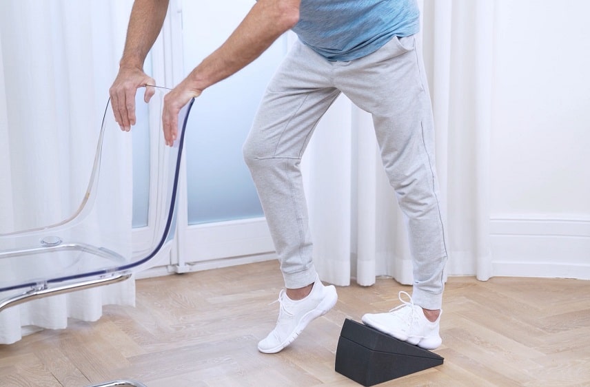 Zu sehen ist der Rumpf-Bein-Bereich eines Mannes in türkisem Oberteil und grauer Jogginghose, der an einem Stuhl lehnt und mit dem linken Bein auf dem Knieretter von Liebscher & Bracht eine Dehnübung für die Wade durchführt. Mit dem rechten Bein steht er auf der Fußspitze.