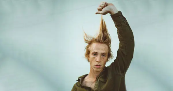 Ein junger Mann in einem grünen Cordhemd zieht seine rot-blonden Haare mit einer Hand nach oben.