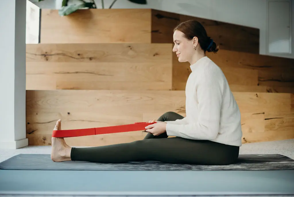 Eine Frau sitzt mit einem ausgestreckten und einem angewinkelten Bein aufrecht auf einer Übungsmatte. Mit einer am au Fuß des ausgestreckten Beines befestigten Übungsschlaufe zieht sie sich leicht nach vorne, um eine Dehnung auszulösen.