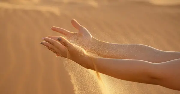 Zu sehen sind zwei lang nach vorne ausgestreckte nackte Arme. Vom Handrücken über das Handgelenk ist ganz leicht ein Henna-Tattoo und die Fingernägel sind in bläulichen Tönen lackiert. Zwischen den Fingern rinnt Sand herunter.