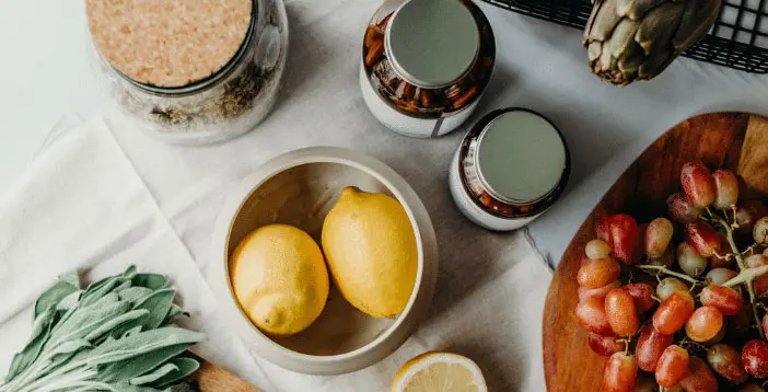 Auf einem Tisch liegen in einer Schüssel 2 Zitronen, ein Holzbrett mit Weintrauben, Rucola, eine Artischocke