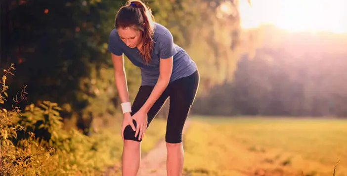 Eine Frau steht in Sportkleidung auf einem kleinen Feldweg und hält mit beiden Händen ihr Knie fest.