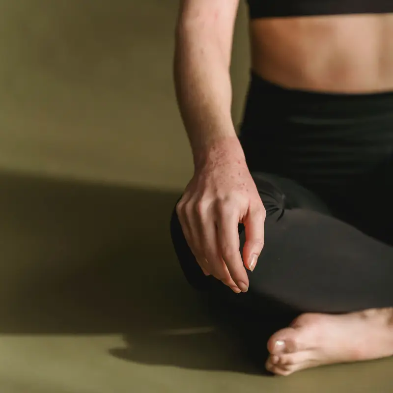 Eine Frau in schwarzer Sporthose und schwarzem Oberteil sitzt im Schneidersitz auf dem Boden. Es ist nur die rechte Hälfte ihres Unterkörpers zu sehen. Die rechte Hand hat sie auf das rechte Knie gelegt.