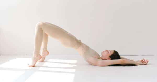 Eine Frau mit braunem Haar im cremefarbenen Trainingsdress liegt in einem weißen Raum auf einer weißen Trainingsmatte und führt eine Brücke durch. Dabei stellt sie sich auf die Zehen und drückt so den Unterkörper nach oben. Die Arme hat sie hinter dem Kopf abgelegt.
