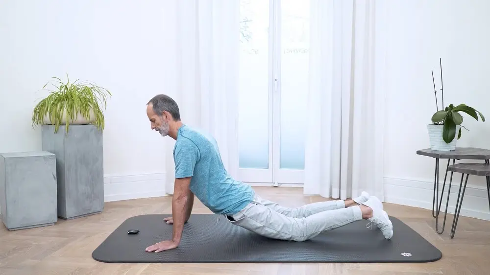 Zu sehen ist ein Mann mit türkisem Shirt, grauer Jogginghose und weißen Turnschuhen der sich mit den Händen auf einer grauen Übungsmatte abstützt und seine Leiste auf die Matte absinken lässt. Er führt auf diese Weise eine Dehnübung für Hüftbeuger gegen Rückenschmerzen im unteren Rückenbereich durch.