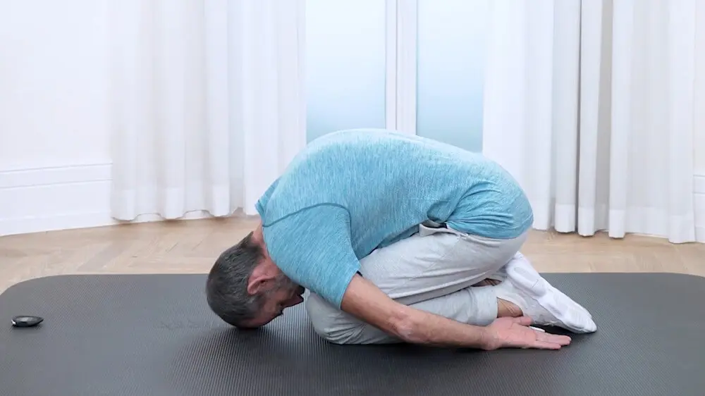 Zu sehen ist ein Mann mit türkisem Shirt, grauer Jogginghose und weißen Turnschuhen, der auf einer grauen Übungsmatte kniet und sich so nach vorne beugt, dass sein Rücken einen Rundrücken bildet. Auf diese Weise gleicht er die Dehnung des unteren Rückens aus.