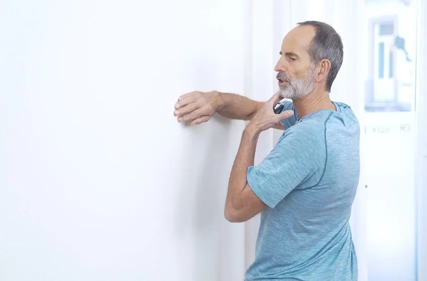 Zu sehen ist ein Mann in türkisem Oberteil, der vor einer Wand steht und den Oberkörper nach links dreht. Mit der rechten Hand fasst er an die Wand, die linke Hand zieht den Kopf nach links. Auf diese Weise führt er eine Rotationsübung zur Dehnung der Hüftbeuger durch.