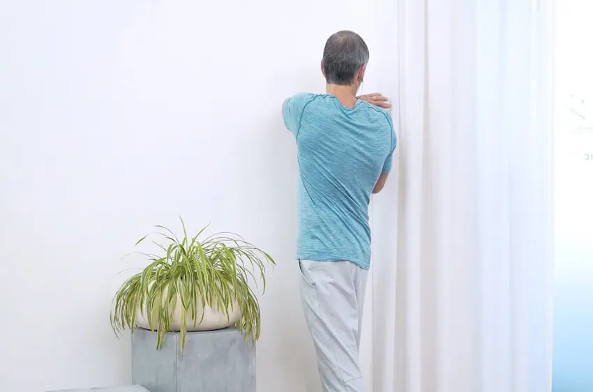 Ein Mann in türkisem Oberteil und grauer Trainingshose steht vor einer Wand. Er befindet sich in einer Rotationsübung, der Oberkörper ist nach rechts gedreht. Links neben ihm steht eine grüne Topfpflanze.