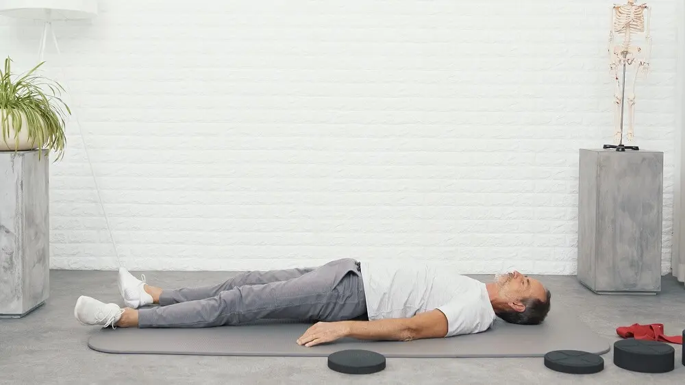 Ein Mann in weißem Shirt und grauer Trainingshose liegt auf einer grauen Trainingsmatte auf dem Rückenretter von Liebscher & Bracht. Andere Teile des Rückenretters sowie eine rote Übungsschlaufe von Liebscher & Bracht liegen neben ihm.
