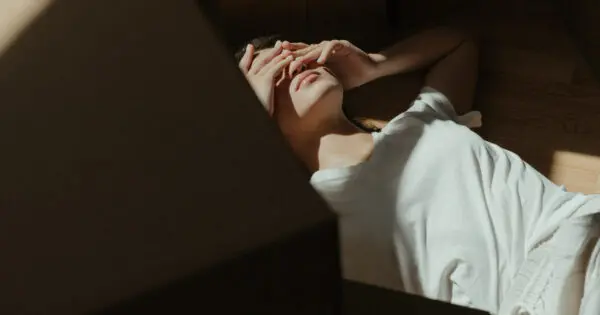 Eine Frau liegt auf dem Boden in der Sonne und um sie herum sind Umzugskisten verteilt. Sie hält sich die Hände vor die Augen und sieht abgeschlagen aus