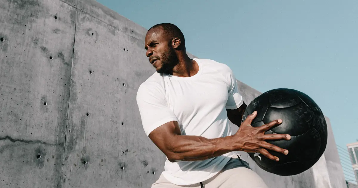 Ein durchtrainierter Mann ist in weißem Sport-Outfit gekleidet. Er hält einen schwarzen Medizinball mit beiden Händen seitlich am Körper und ist leicht gedreht. Er setzt zum Wurf an.