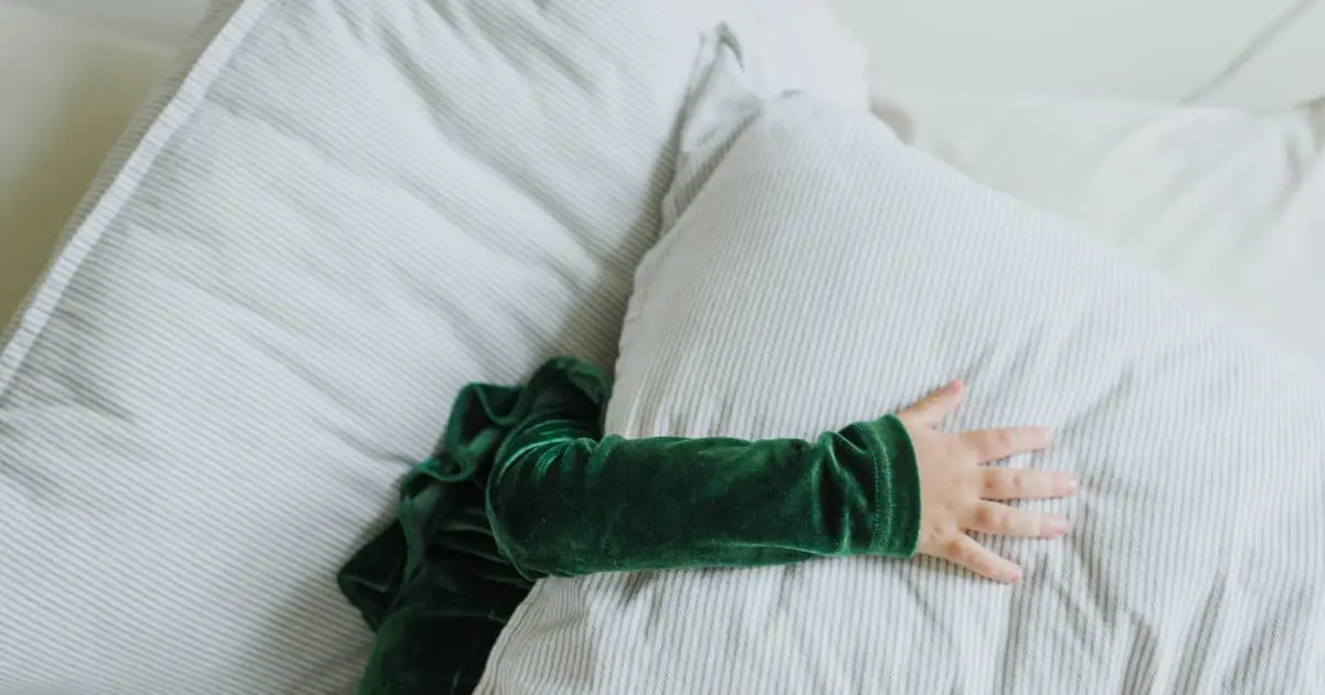 Ein Kind liegt auf einem Bett, es sind nur ein großes Kissen sowie ein Arm in grünem Pullover zu sehen.