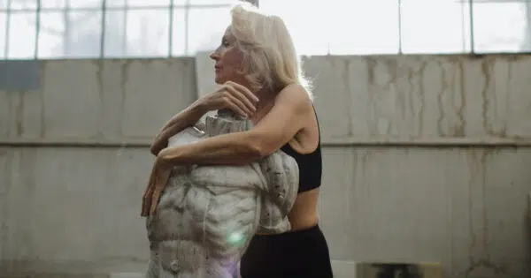 Eine hübsche ältere Frau steht in einem schwarzen Yoga-Outfit in einer großen Fabrikhalle. Sie umarmt mit ihren Armen einen steinernen Torso und ist im Profil dargestellt.