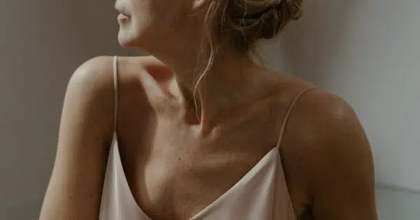 Eine ältere Frau in einem rosa Satin-Spaghetti-Top schaut gedankenvoll zur Seite. Man sieht ihre nackten Oberarme, die einen Bizeps erahnen lassen.