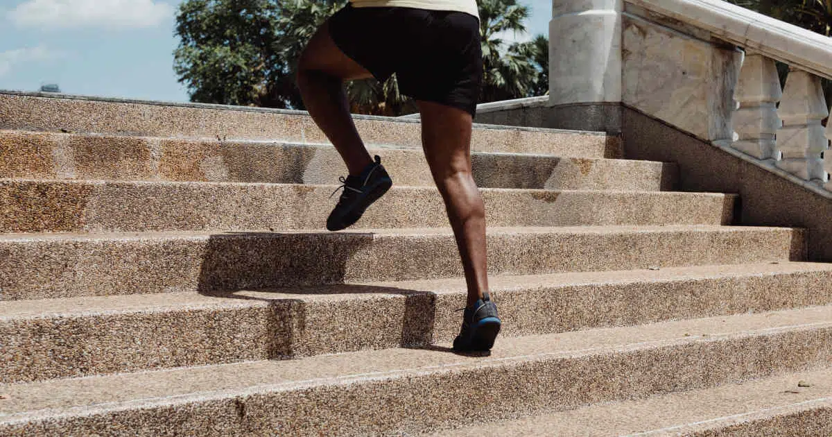 Ein Mann mit dunkler Hautfarbe und kurzen Hosen joggt eine breite Treppe hinauf. Die Sonne scheint und der Körper wirft einen Schatten auf die helle Treppe. Der Mann trägt schwarze Shorts und schwarze Schuhe. Im Hintergrund sind Palmen zu sehen.