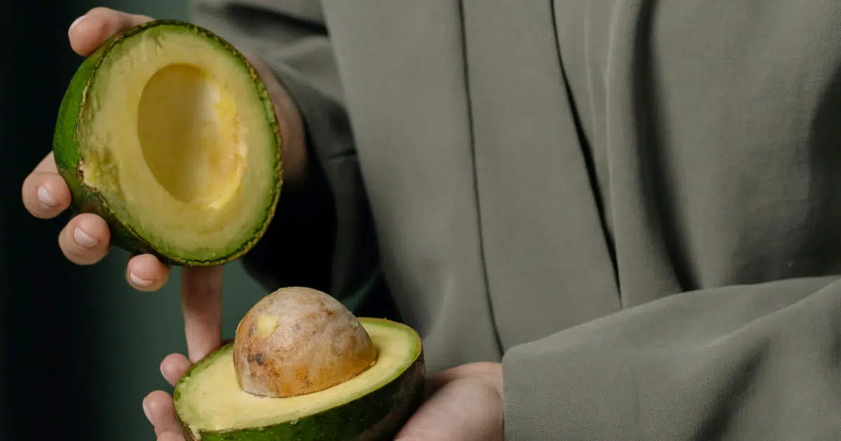 Eine junge Frau hält eine aufgeschnittene Avocado in ihren Händen. Sie trägt einen kaki-grünen Mantel.