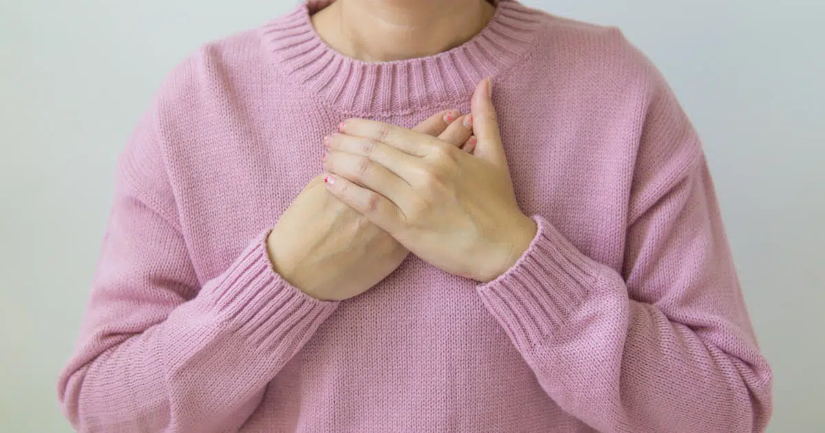 Das Bild zeigt den Rumpf einer Frau. Sie trägt einen rosafarbenen Pullover und legt ihre Hände gekreuzt auf das Brustbein.