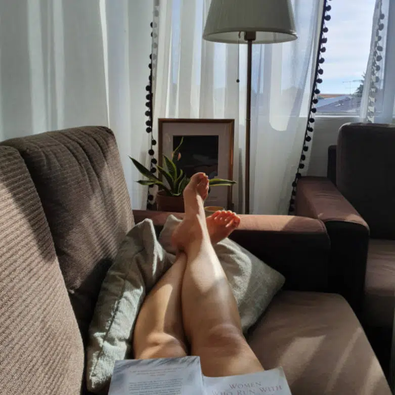 Auf einem braunen Sofa mit Blick zum Fenster liegt eine Frau. Sie hat ihre Beine ausgestreckt und die Füße hochgelegt. Auf ihrem Schoß liegt ein aufgeklapptes Buch.