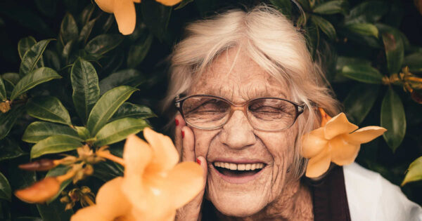 Eine ältere Dame mit grauen Haaren und Brille steht in einem Busch mit orangenen Blüten. Sie sieht glücklich aus und hält sich mit der rechten Hand an die rechte Gesichtshälfte